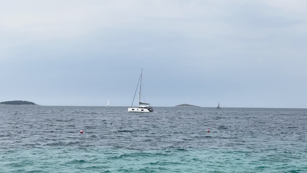 A white sailboat calmly drifts on the open sea with a few distant boats on the horizon. Overcast sky creates a peaceful maritime vacation mood