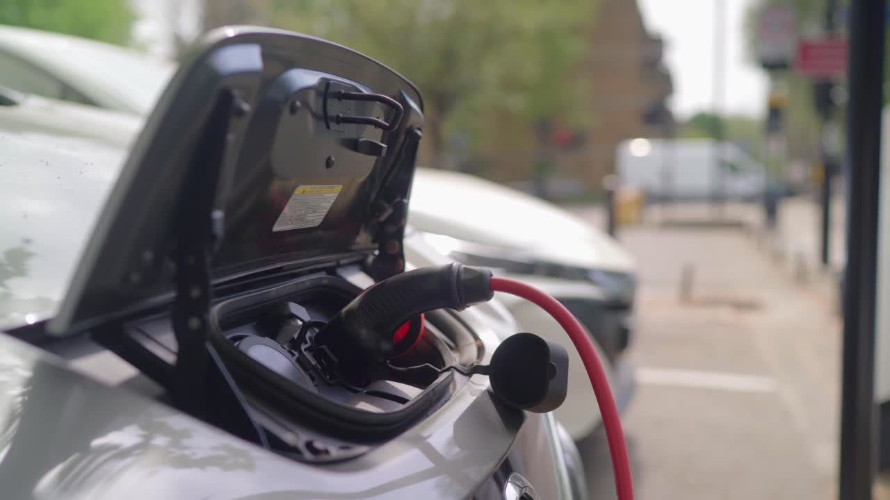 An electric car is parked near the charging station and being charged with a red cable as the hood in the front of the car is open during daytime.