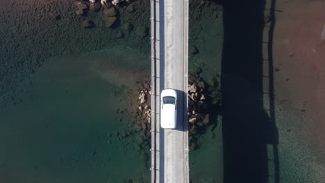 White-car-parked-in-middle-of-bridge-with-calm-blue-water-on-sunny-day,-aerial