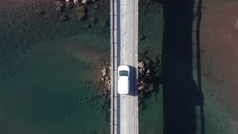 white car parked in middle of bridge with calm blue water on sunny day, aerial