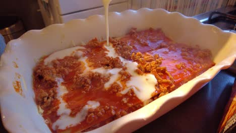 se vierte salsa bechamel sobre boloñesa caliente dentro de un plato de lasaña con queso rociado después