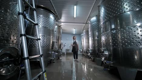 a happy successful farmer or winemaker is walking in the middle of wine tanks and checking a fermentation grade of high quality wine production in a modern winemaking facility or winery.