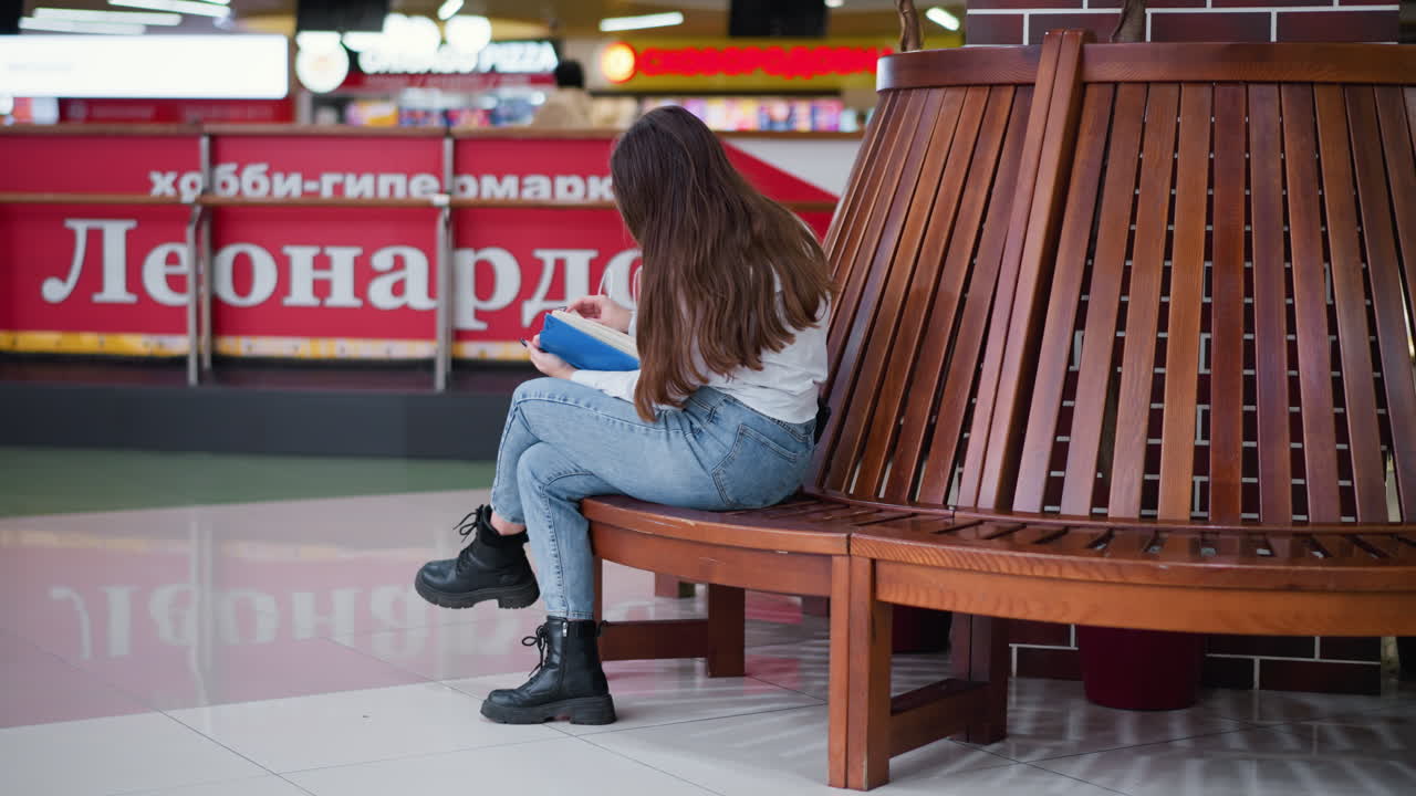 vista trasera de una mujer sentada en un banco de madera, con las piernas cruzadas, leyendo un libro, el cabello largo fluyendo, compradores visibles en el fondo, entorno vibrante del centro comercial