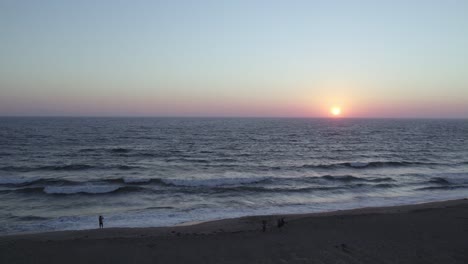 Gorgeous-Orange-and-Pink-Sunset-in-Oxnard,-California---4K-Drone-Over-the-Ocean-with-a-Pull-Back-to-Reveal-Beach-Goers-Watching-the-Sunset