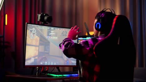 vista trasera de un streamer asiático que se estira mientras juega un juego a través de la red en una computadora personal. videojuego de transmisión en vivo, escritorio iluminado por luz de tira de led rgb