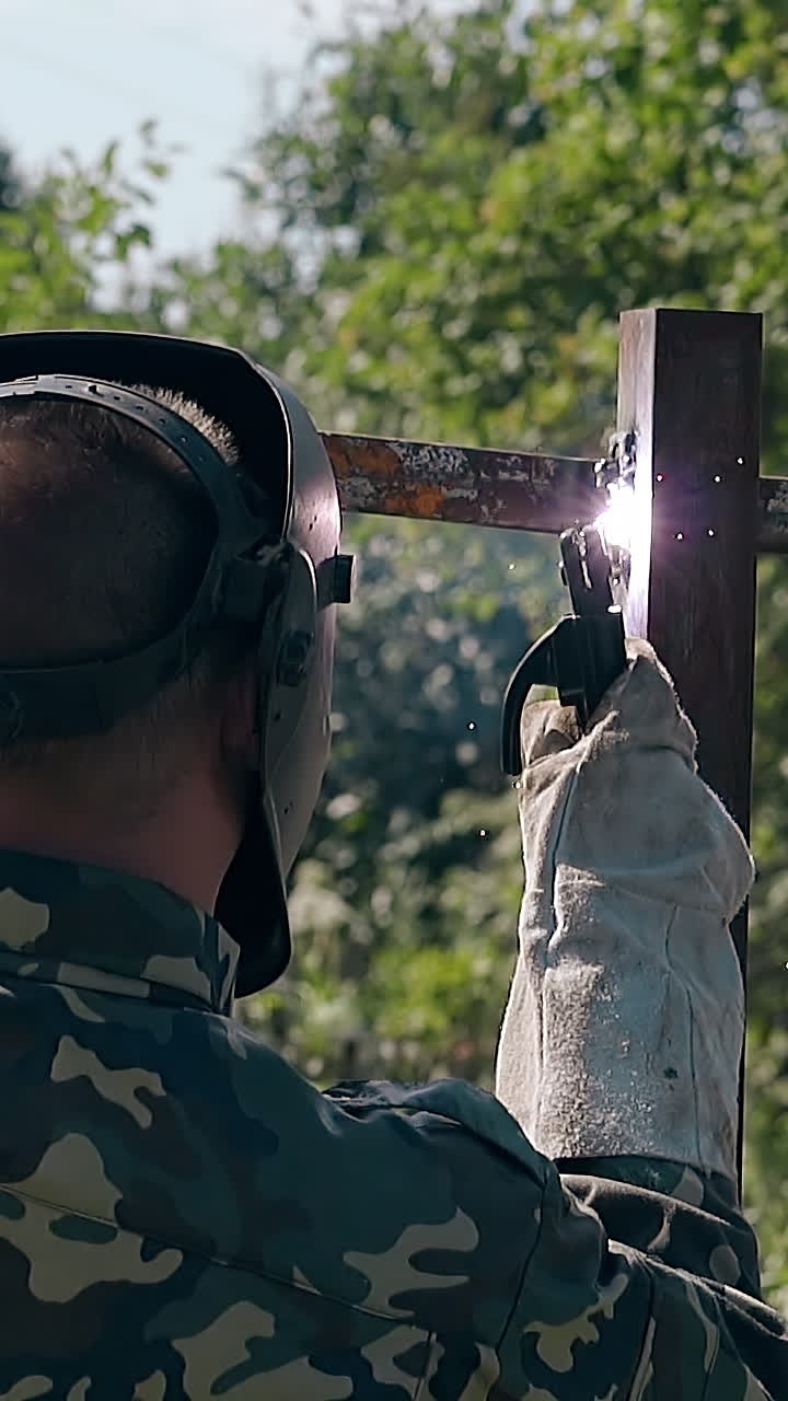 handyman in camouflage and work gloves welds plank of metal fence in garden slow motion backside view