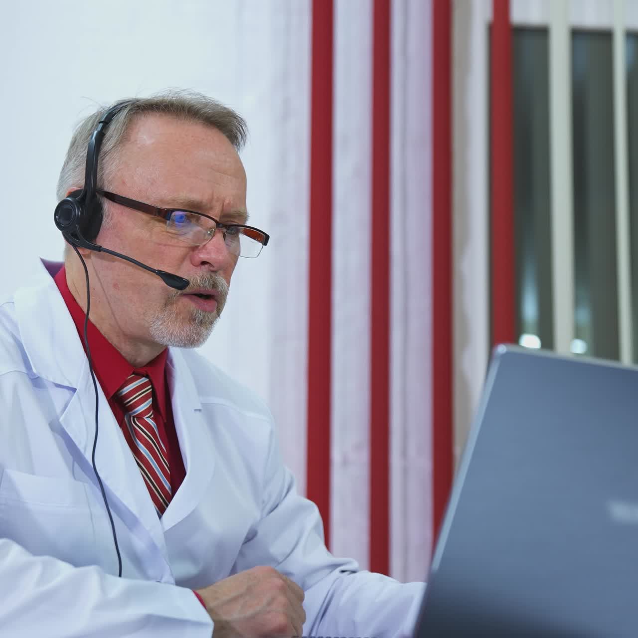 Telemedicina. Anziano medico ascolta un paziente dal laptop. Medico uomo con cuffie che effettua una consulenza tramite videochiamata online con un paziente in clinica