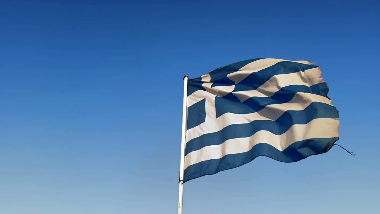 Worn Greek flag close up, waving against clear blue sky, symbolizing resilience