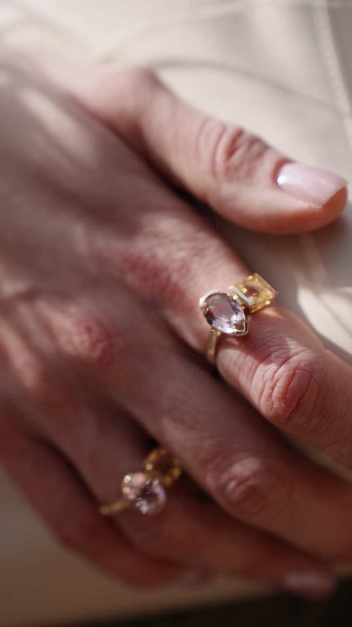 A close-up of hand adorned with multiple elegant colorful gemstones shiny rings resting on on a stylish bag outdoors in sunshine