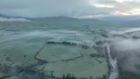 flying over meandering river towards mist shrouded patchwork fields at dawn in winter