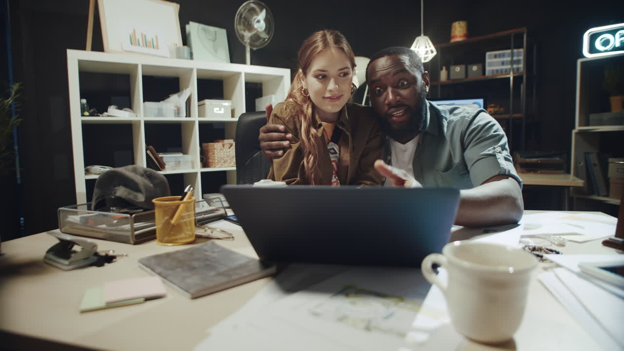 un hombre afro coqueteando abrazando a una hermosa mujer cerca de una computadora portátil en una oficina hipster.
