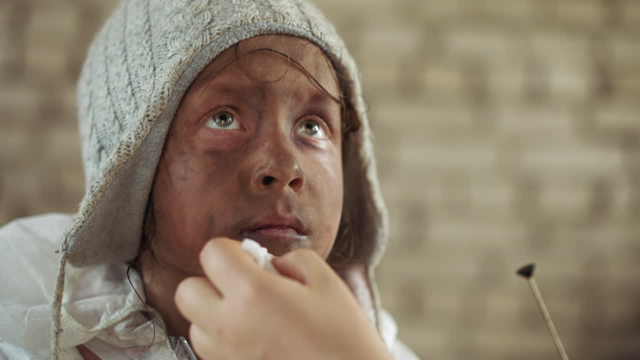 Close up of dirty child in knitted beanie being gently wiped clean by helping hand, showing emotional expression of fatigue and vulnerability inside shelter with blurred wall in background