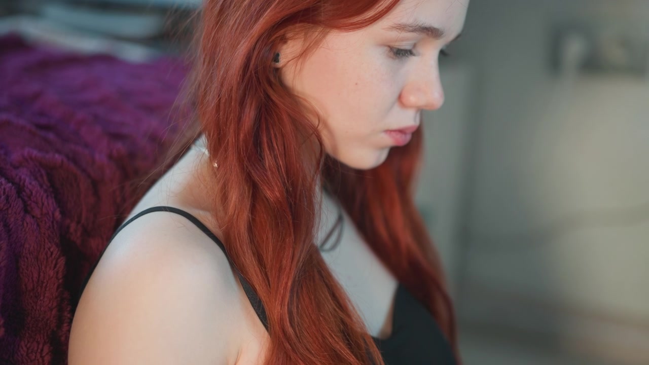 Young caucasian redhead reading tarot cards in bedroom, apprentice diviner flips guidebook, home reader examines card spread, intuitive consultant holds talisman and studies handwritten note, soft