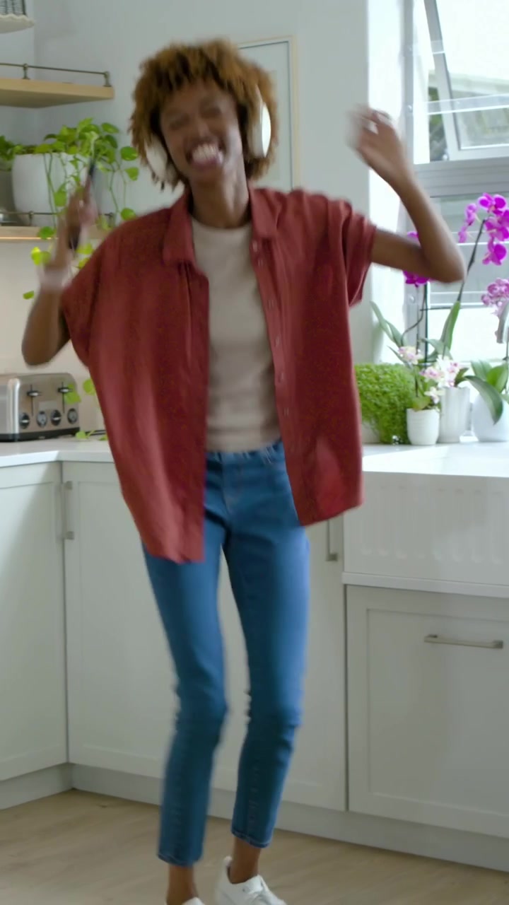 Vertical video: Dancing woman with headphones and red shirt moving in kitchen, phone playing music