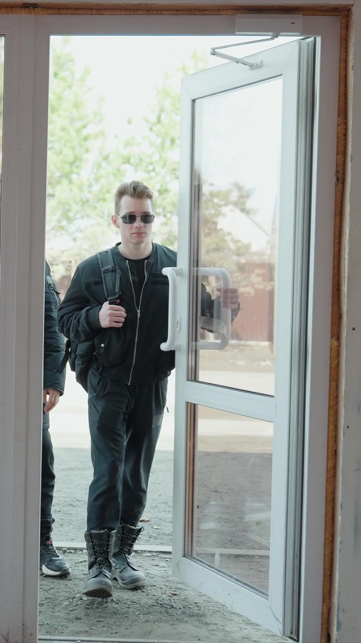 Young man in dark clothing walks towards glass door adjusting sunglasses. Another man stands inside doorway holding it open. Scene transitions between indoor views