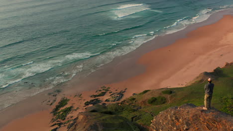 Aerial:-A-man-standing-on-a-viewpoint-watching-the-surfers-in-Portugal