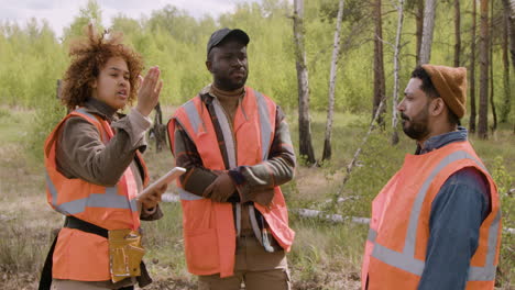 ativista afro-americana segurando um tablet e conversando com seus colegas de trabalho na floresta enquanto eles decidem onde plantar árvores