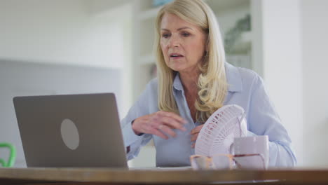 menopausal mature woman having hot flush at home cooling herself with fan connected to laptop