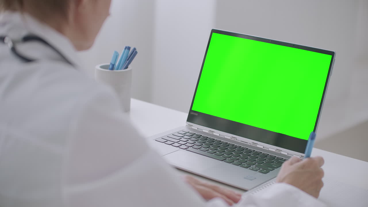 la doctora está aprendiendo en línea mirando la pantalla verde de la computadora portátil para la tecnología de clave de croma escribiendo notas en papel y asintiendo con la cabeza