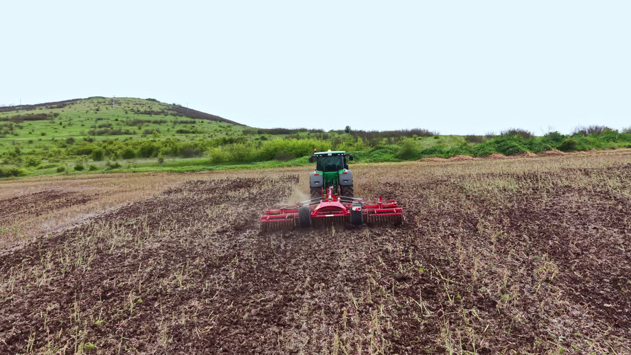 Agricultor utiliza trator para lavrar o solo em um campo em um dia ensolarado em uma área rural cercada por colinas.