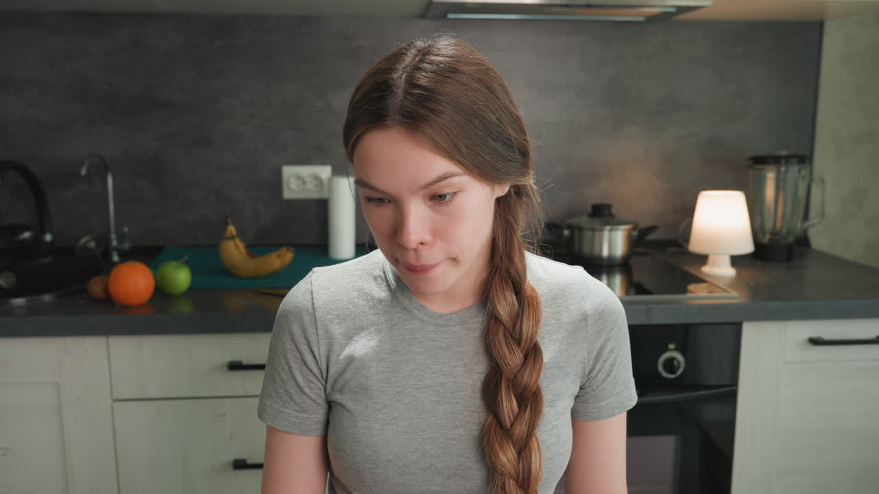 Donna bianca incinta che guarda in basso in cucina, con capelli intrecciati, maglietta grigia, frutta e banana sul bancone, una sottile espressione di disagio che suggerisce nausea o avversione al cibo, un momento autentico di una futura mamma, domestico.