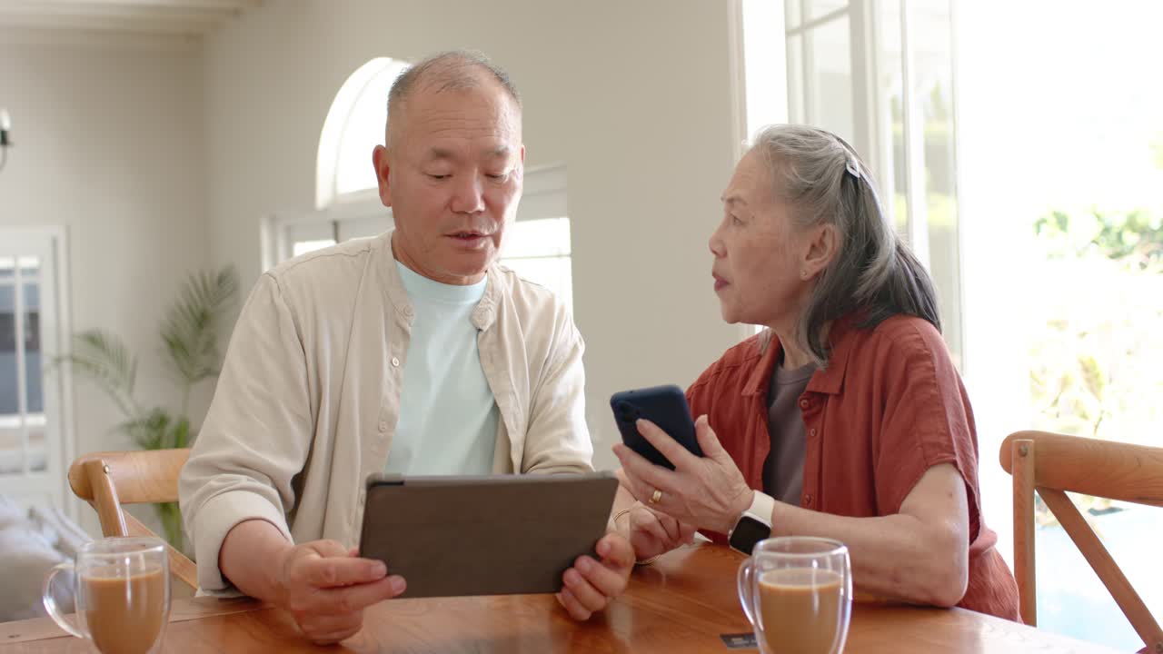 アジア人カップルがキッチンテーブルに座り、女性がスマートフォンを見せ、男性はタブレットを操作して情報を確認している