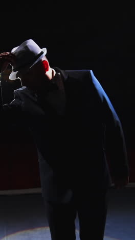male actor performing in the theater. funny man putting on white hat and began dancing on stage in dark light. view from the stage. vertical video