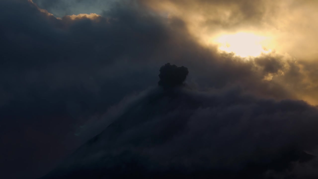 Volcán activo arrojando ceniza y humo a un cielo dramático con nubes resplandecientes durante un atardecer.