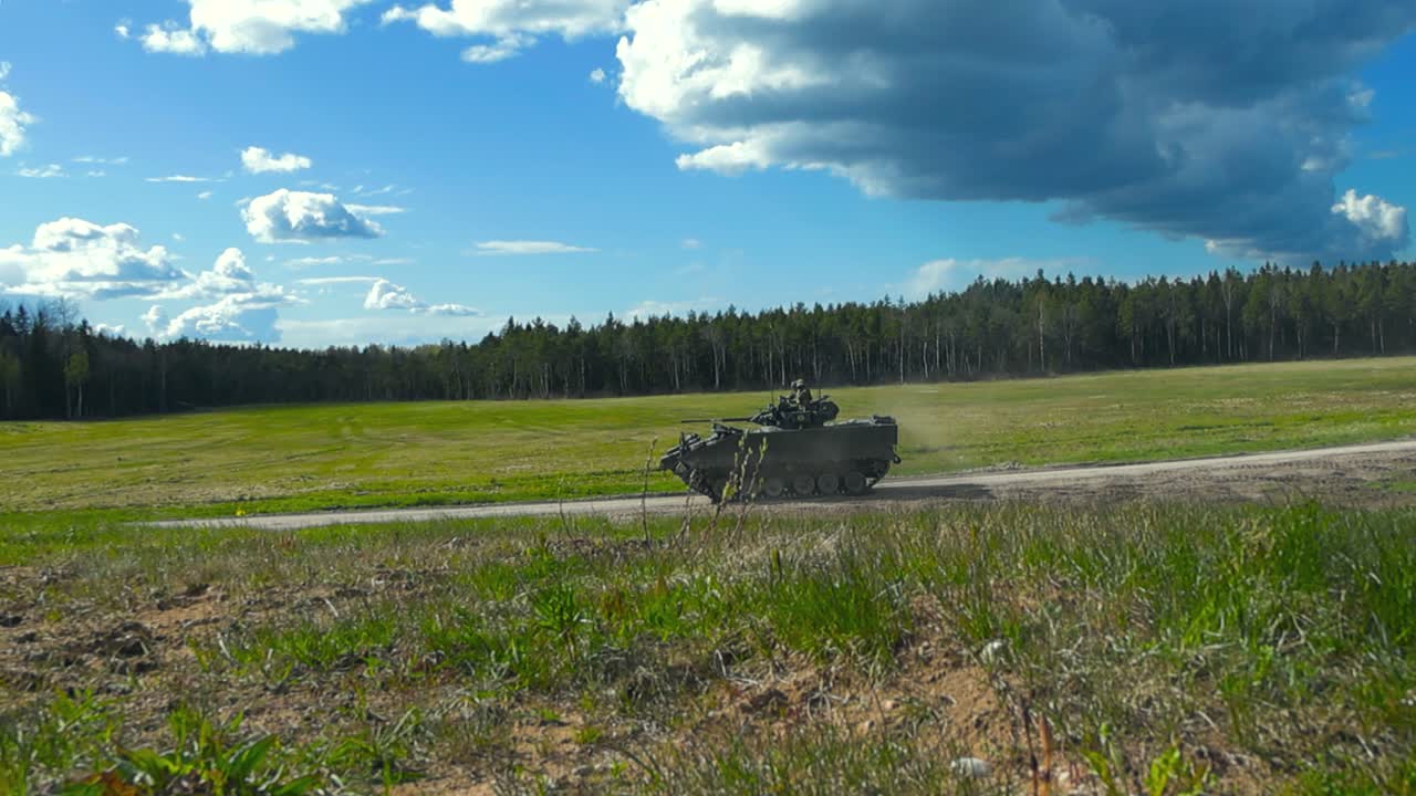 Gorgeous rare footage of a British Warrior FV510 tank or armoured personnel carrier driving from a grassy green summer field to a gravel road and moving ahead in slow motion with dust cloud behind it.