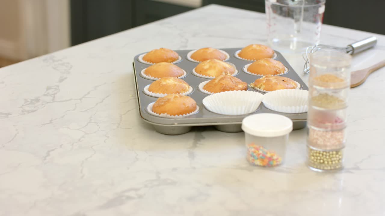 Camera che inquadra dodici cupcake dorati su un piano di lavoro con granelli e una frusta in attesa di essere decorati