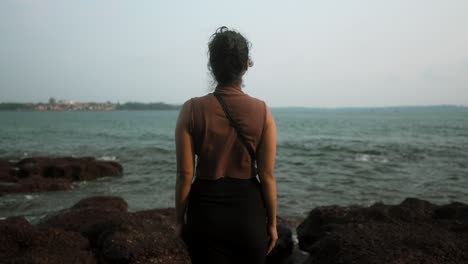 Woman-stands-on-rocky-shore-looking-at-the-sea-during-a-peaceful-evening