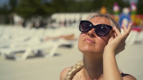 close-up travelling blonde girl is sitting on deck chair on the beach in sunglasses her hair is braided