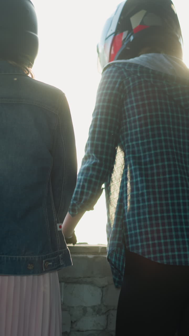 창문 앞에 서 있는 두 여성의 뒷면, 한 명은 검은색 레과 스니커를 입고, 다른 한 명은 플리티드 스커트와 젠임 재을 입고, 둘 다 차분하게 보입니다.