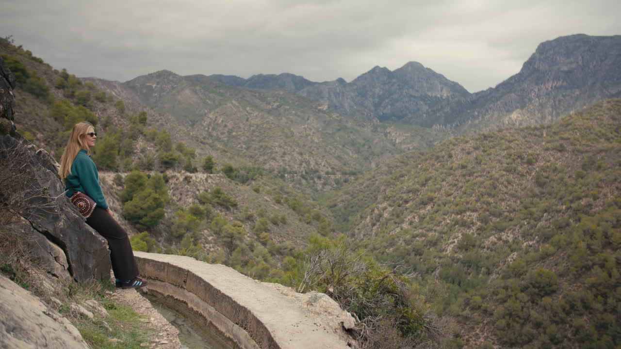 Donna seduta tranquillamente su un pendio roccioso accanto all'acquedotto, con vista su una vasta valle montana verde. Filmato su un sentiero remoto nel Sud della Spagna.