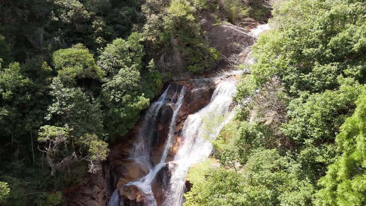 A beautiful aerial shot pushing in on a powerful waterfall. The water cascades over large rocks, surrounded by a lush, green forest in a remote Japanese mountain landscape.
