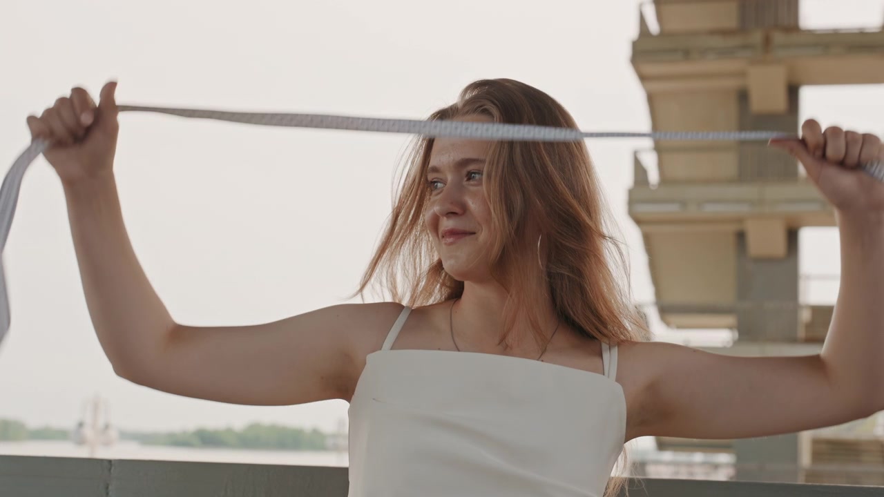 Woman in white dress holds and measures herself with tape measure standing on outdoor bridge or balcony. Her hair moves naturally as woman looks around scene which includes soft daylight
