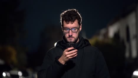 Young-Man-stands-in-cold-winter-night-in-german-suburb-around-munich-and-is-scrolling-on-his-phone,-using-technology