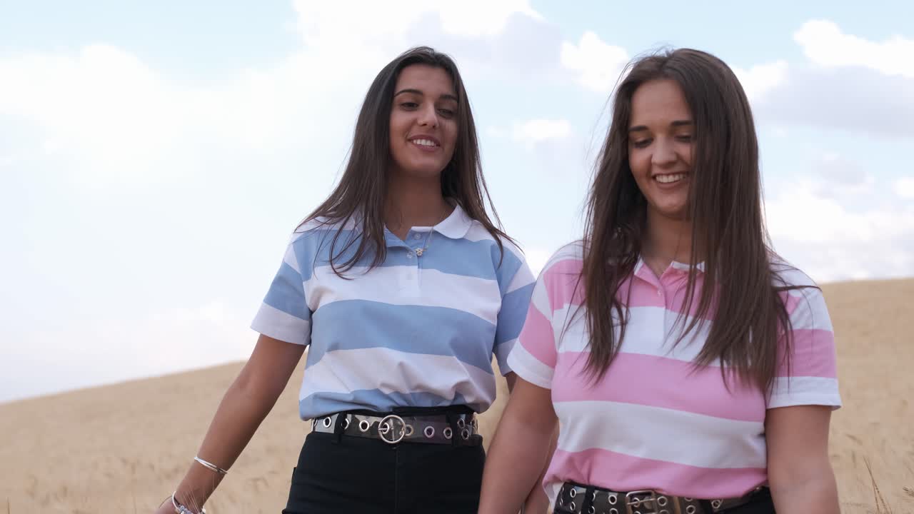 Front view of two female friends holding hands and smiling while walking in a wheat field together.