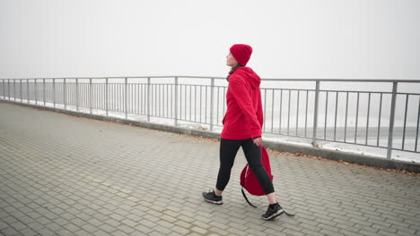 business woman holds backpack as rope drags along ground, walking on interlocked path near iron railing with scattered dry foliage and moving car visible below bridge in foggy atmosphere