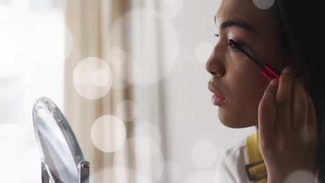 Animation-of-light-spots-over-biracial-woman-applying-mascara