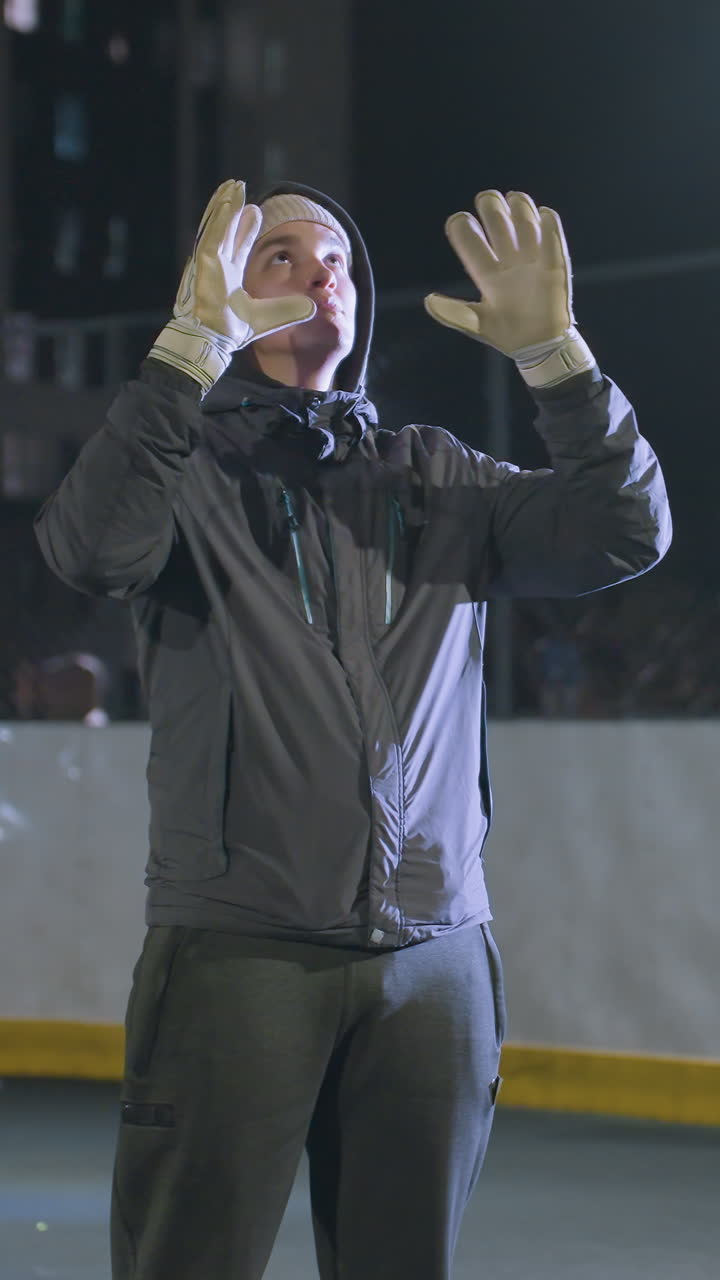 futbolista con guantes lanzando una pelota de fútbol en el aire y atrapándola durante la práctica nocturna, telón de fondo urbano con edificio borroso, efectos de luz bokeh, y tres personas caminando detrás de la valla