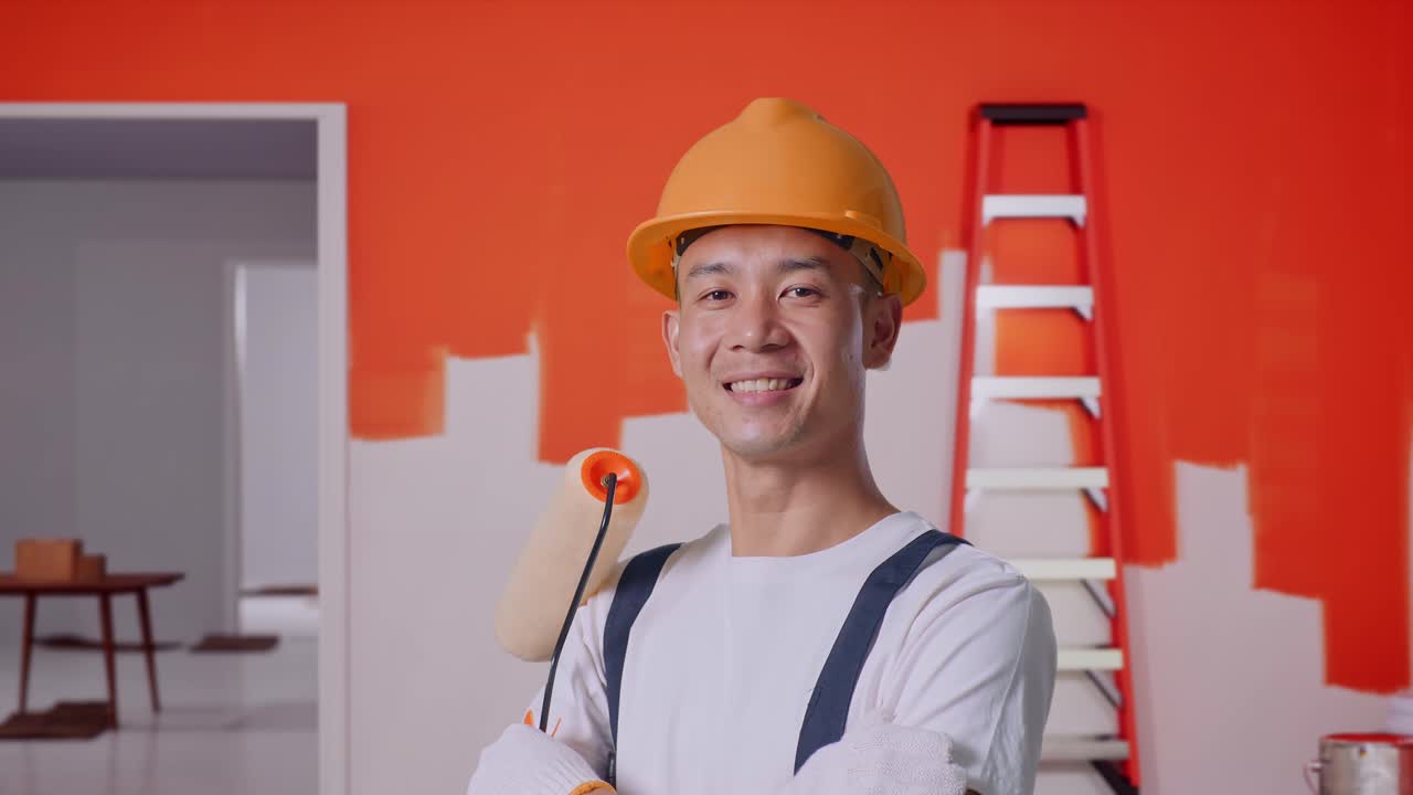 Close Up Side View Of Asian Man Painter Wearing Safety Helmet Smiling And Crossing His Arms While Standing With Painting Wall In Room