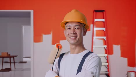 vista lateral cercana de un pintor asiático que usa un casco de seguridad sonriendo y cruzando los brazos mientras está de pie con la pared de pintura en la habitación