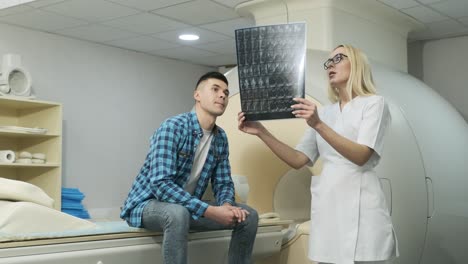 la radióloga explica los resultados de la resonancia magnética para un paciente joven, mostrando la instantánea con imágenes, observando y analizando la resonancia eléctrica en una clínica moderna junto a un escáner de resonancia magnético moderno