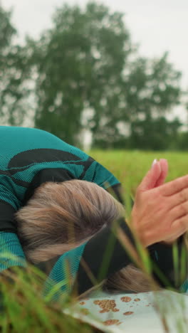 mujer de traje verde y negro sentada en una alfombra de yoga, inclinada hacia adelante con las manos extendidas y la cabeza tocando la alfombra, practicando una postura de yoga al aire libre en un campo de hierba bajo un cielo nublado