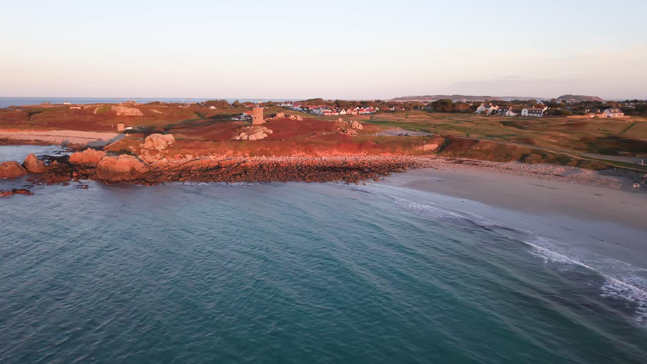 Flight along shoreline of Lancresse Bay Guernsey with calm water,beach and rocks in the stunning colours of the golden hour towards Martello Tower sat on the headland in golden sun
