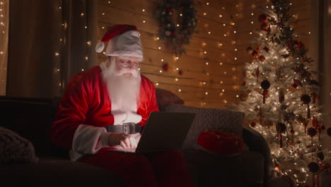 Portrait-of-Santa-Claus-at-home-sitting-on-the-sofa-with-a-laptop-near-the-Christmas-tree-working-with-a-laptop