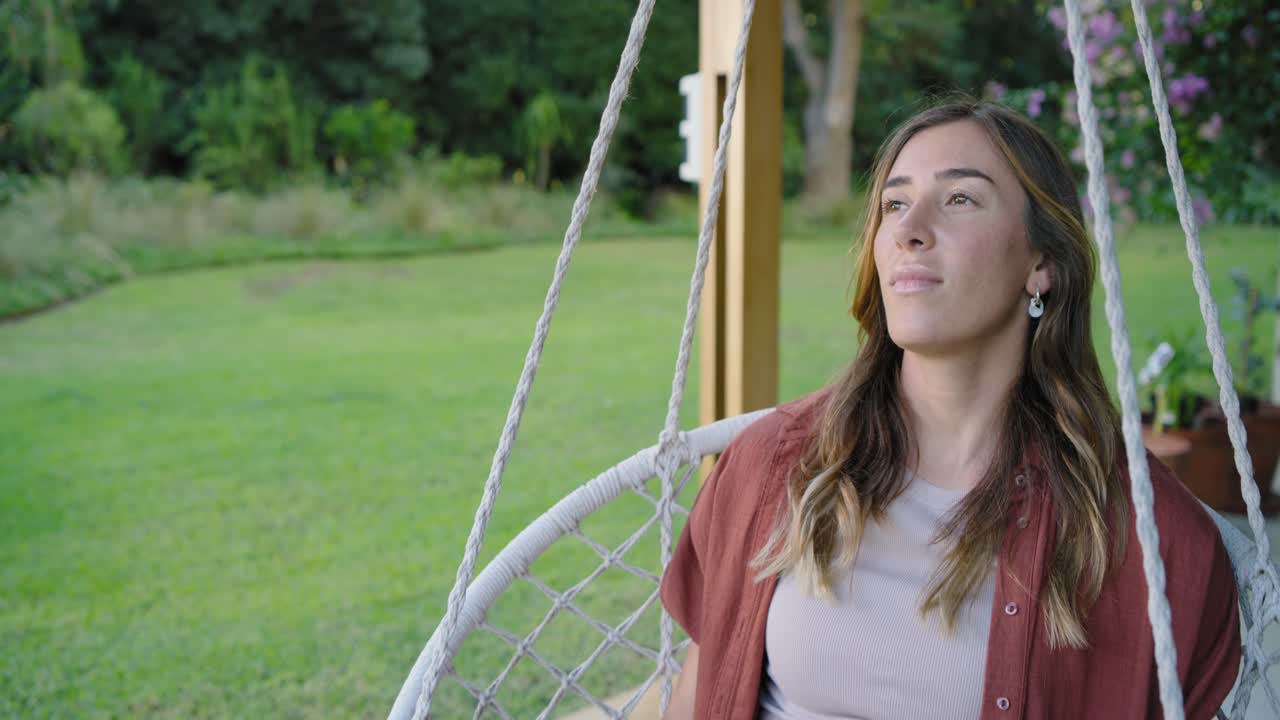 Mulher vestindo camisa cor ferrugem sentada em cadeira de macramê na varanda segurando cordas, olhando para cima, relaxando