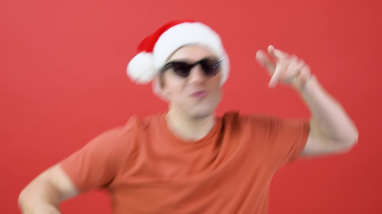 un hombre con sombrero de papá noel y gafas de sol baila agitando los brazos. el tipo con el traje rojo de navidad se encuentra frente a un fondo rojo