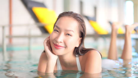 portrait of young happy beautiful asian woman lying in spa center swimming pool and smiling to camera, full frame